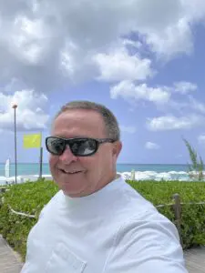 A man in sunglasses and white shirt standing next to the ocean.